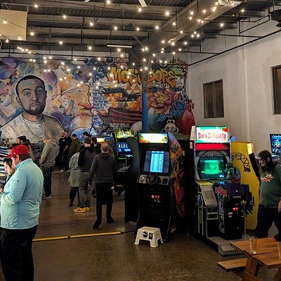 A small group of people play pinball machines at a large warehouse space. In the background is a mural featuring the late rapper Mac Miller.